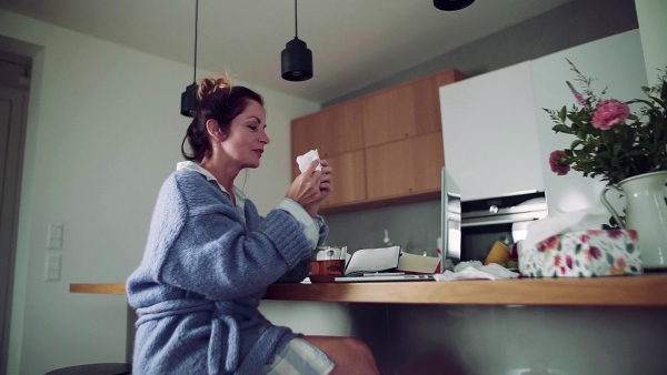 A sick mature woman with laptop sitting indoors, drinking tea when working in home office. Slow motion.