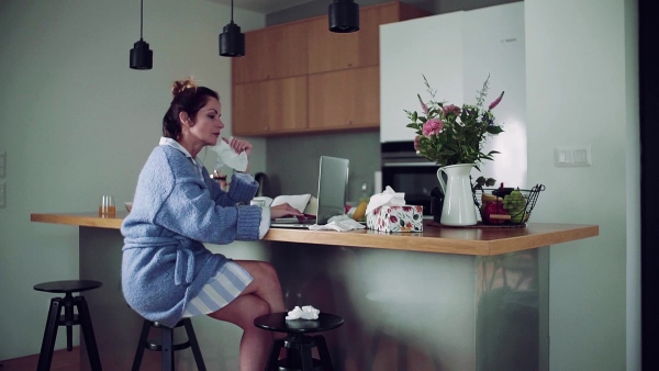 A sick mature woman with laptop sitting indoors, drinking tea when working in home office. Slow motion.