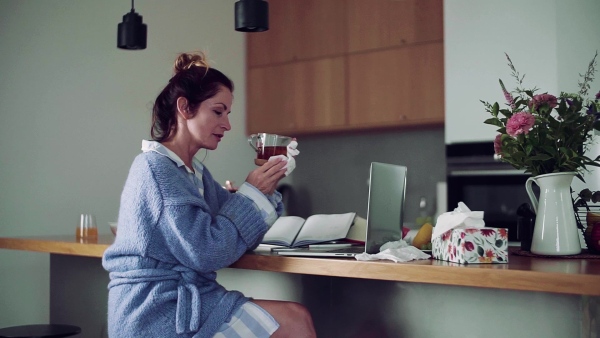 A sick mature woman with laptop sitting indoors, drinking tea when working in home office. Slow motion.