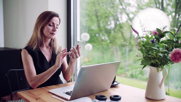 A mature woman with laptop sitting at the table, using smartphone in home office. Slow motion.