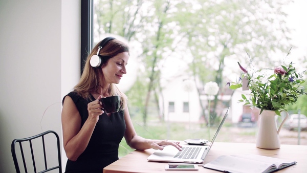 A mature woman with laptop and headphones sitting at the table in home office. Slow motion.