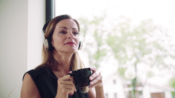 A mature woman with coffee and headphones sitting at the table in home office. Slow motion.