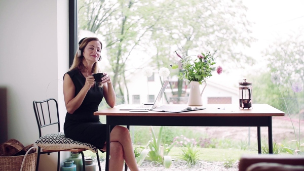 A mature woman with laptop and headphones sitting at the table in home office. Slow motion.