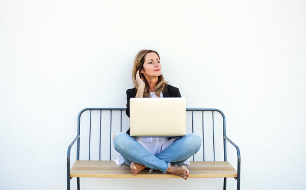 Happy mature woman working in home office outdoors on bench, using laptop.