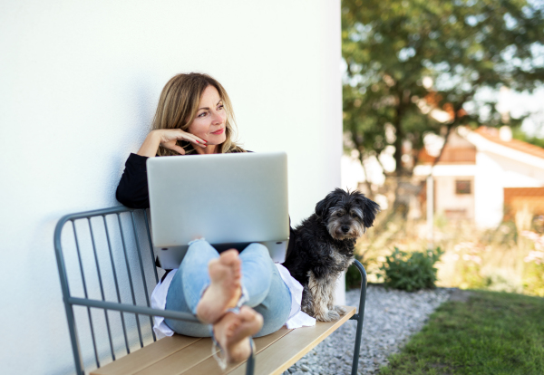 Happy mature woman with dog working in home office outdoors on bench, using laptop.