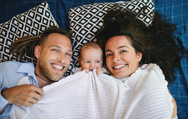 Portrait of young couple with baby daughter resting under blanket, looking at camera.