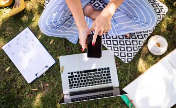 Unrecognizable woman in pajamas working in home office outdoors in garden, using smartphone and laptop.