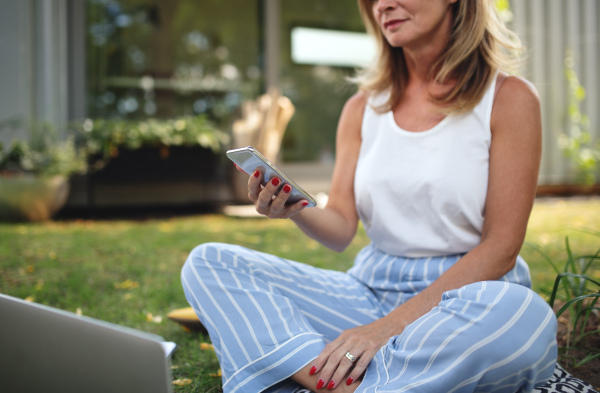 Unrecognizable mature woman in pajamas working in home office outdoors on grass in garden, using smartphone.