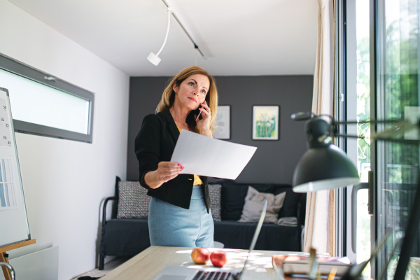 Mature woman working indoors in home office in container house in backyard, using smartphone.