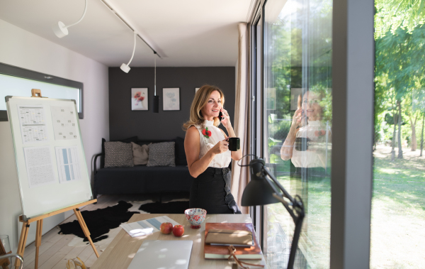 Mature woman working indoors in home office in container house in backyard, using smartphone.