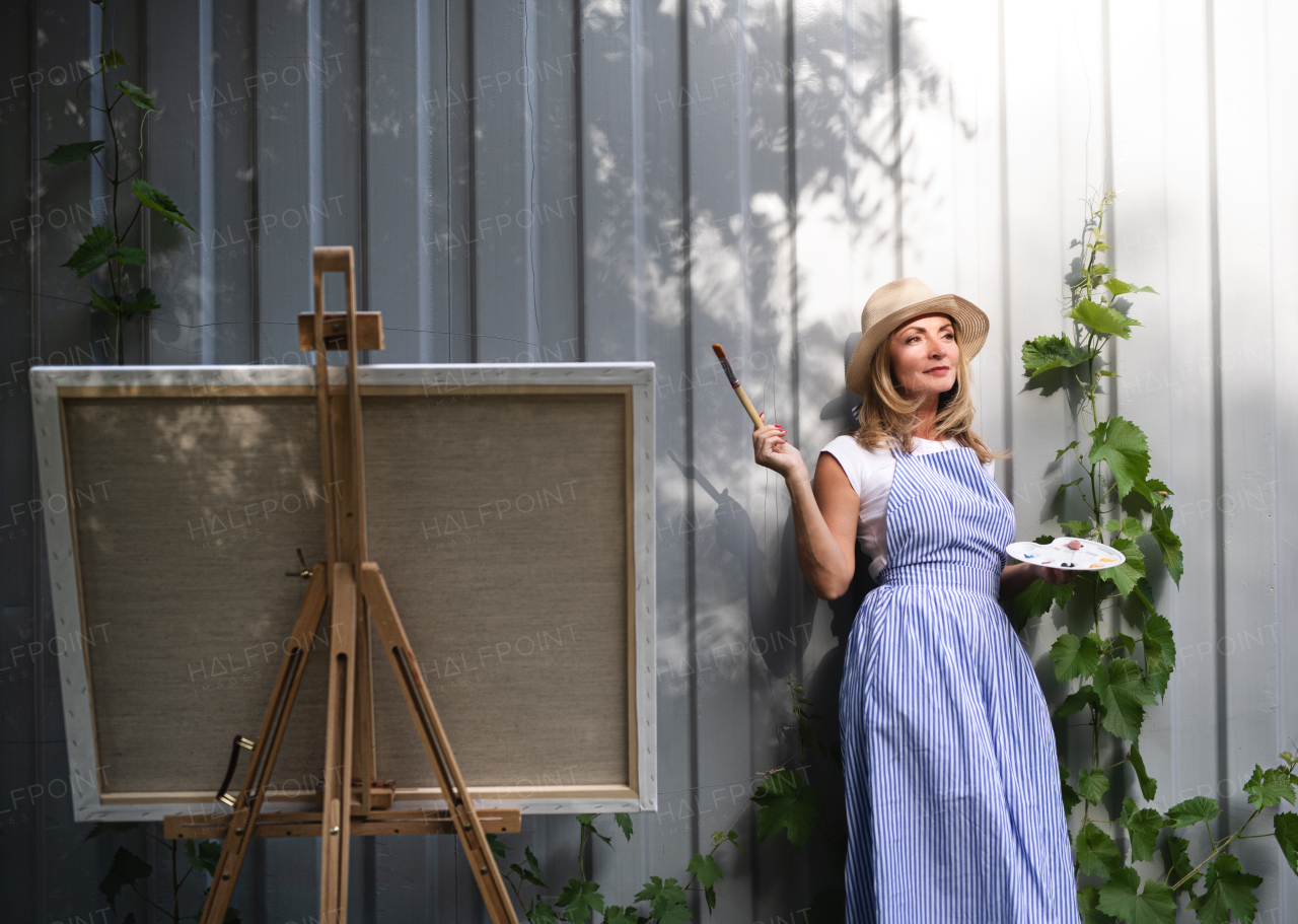 Portrait of happy mature woman with pallete painting outdoors in garden.