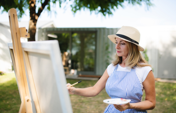 Portrait of happy mature woman with pallete painting outdoors in garden.