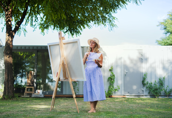 Portrait of happy mature woman with pallete painting outdoors in garden.