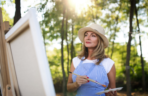 Portrait of happy mature woman with pallete painting outdoors in garden.