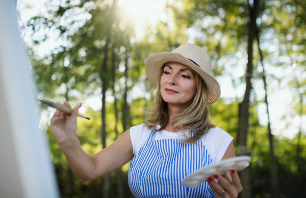 Portrait of happy mature woman with pallete painting outdoors in garden.