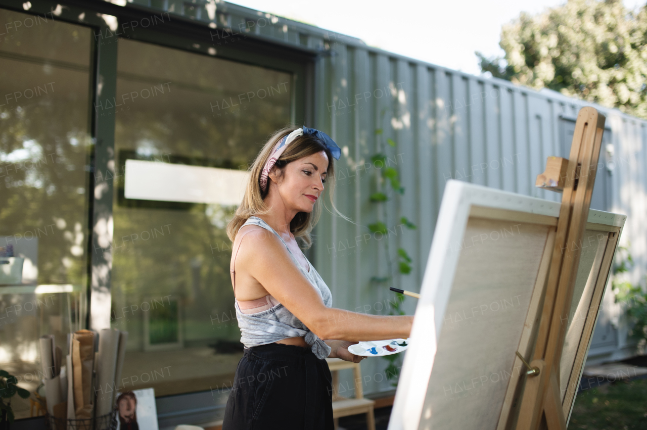 Portrait of happy mature woman with pallete painting outdoors in garden.