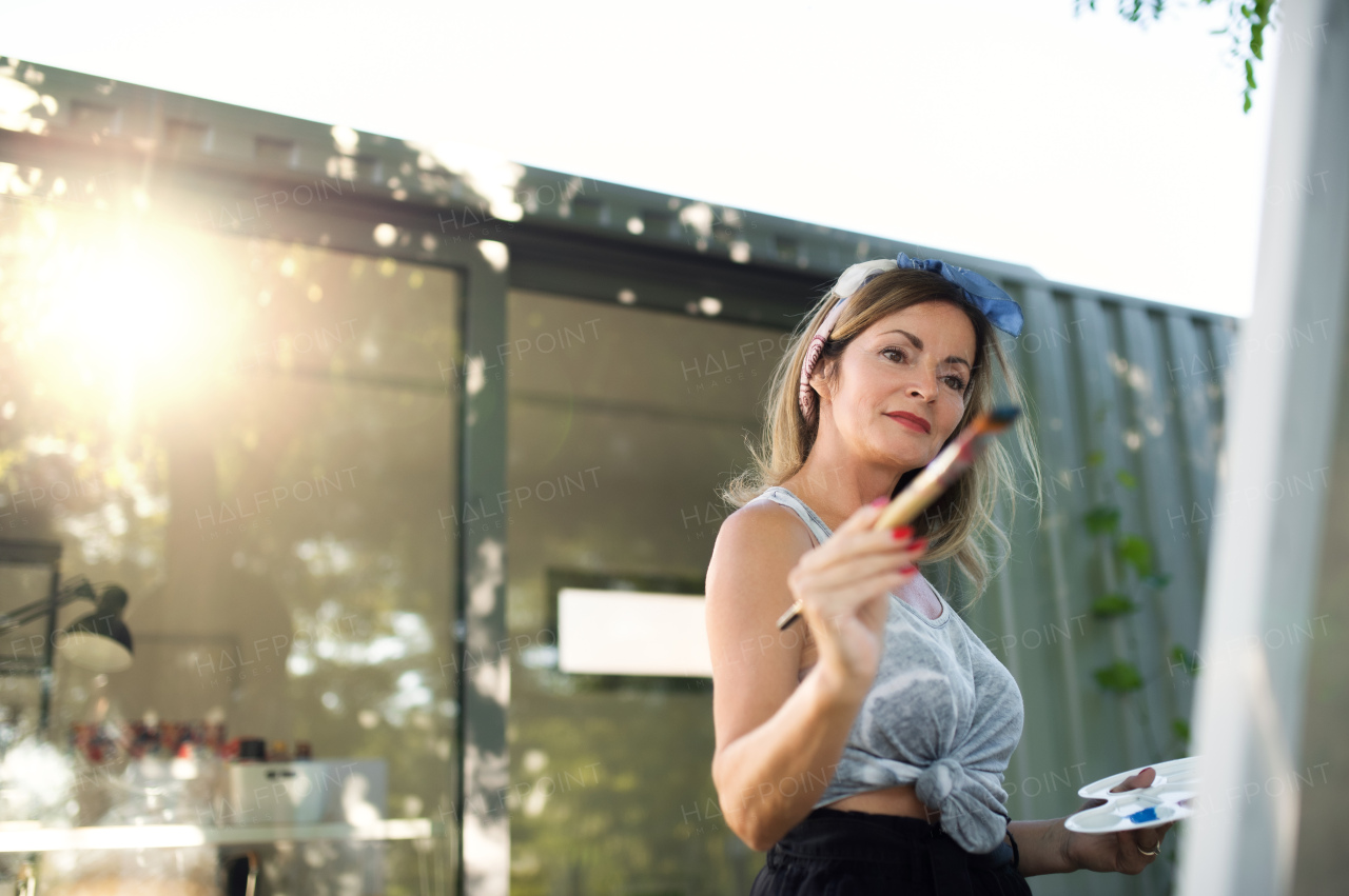 Portrait of happy mature woman with pallete painting outdoors in garden.