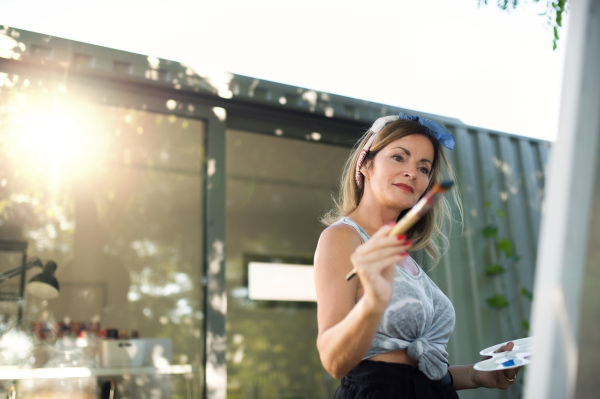 Portrait of happy mature woman with pallete painting outdoors in garden.