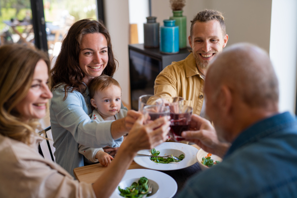 A happy multigeneration family indoors at home eating healthy lunch, clinking glasses.