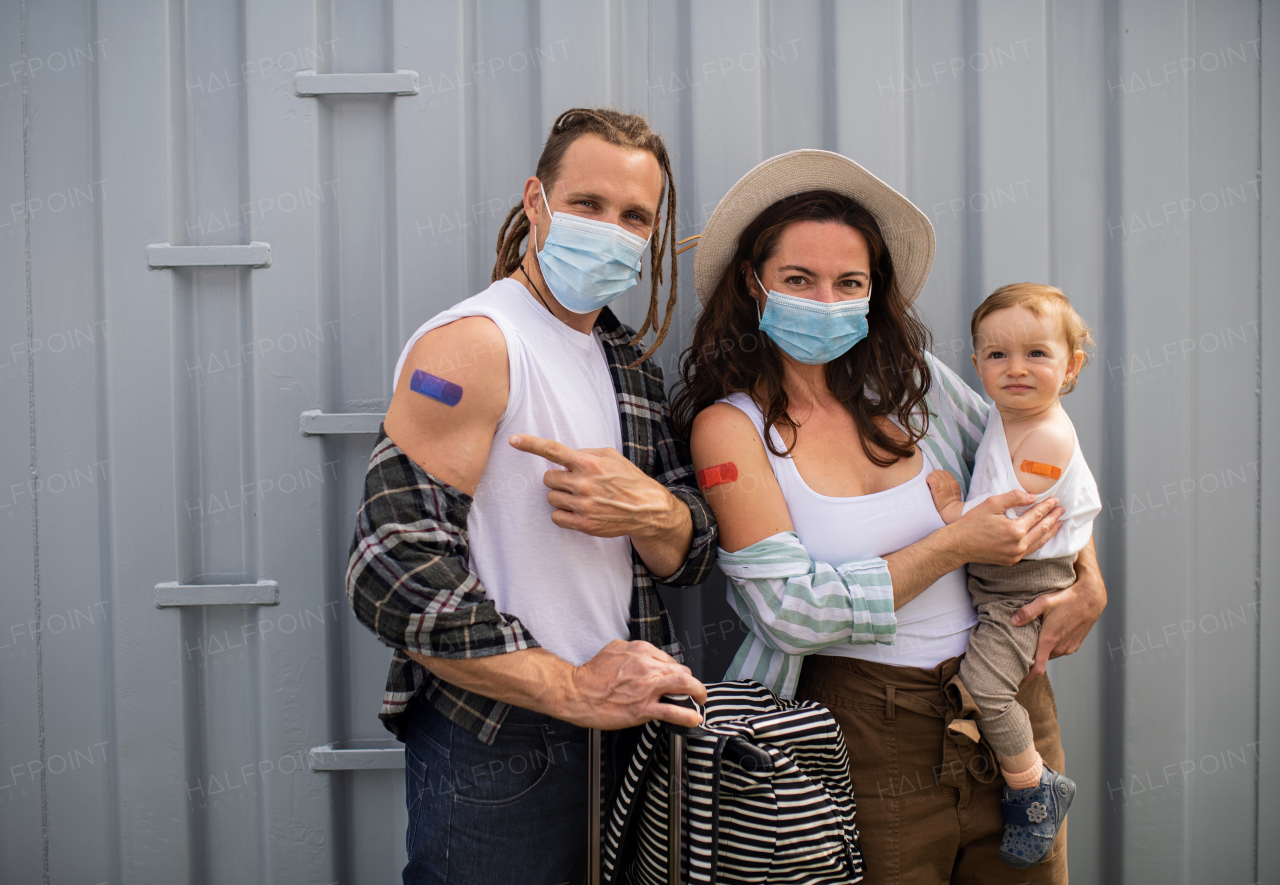 A portrait of young family with small baby after covid-9 vaccination, pointing to plaster on arm and looking at camera.