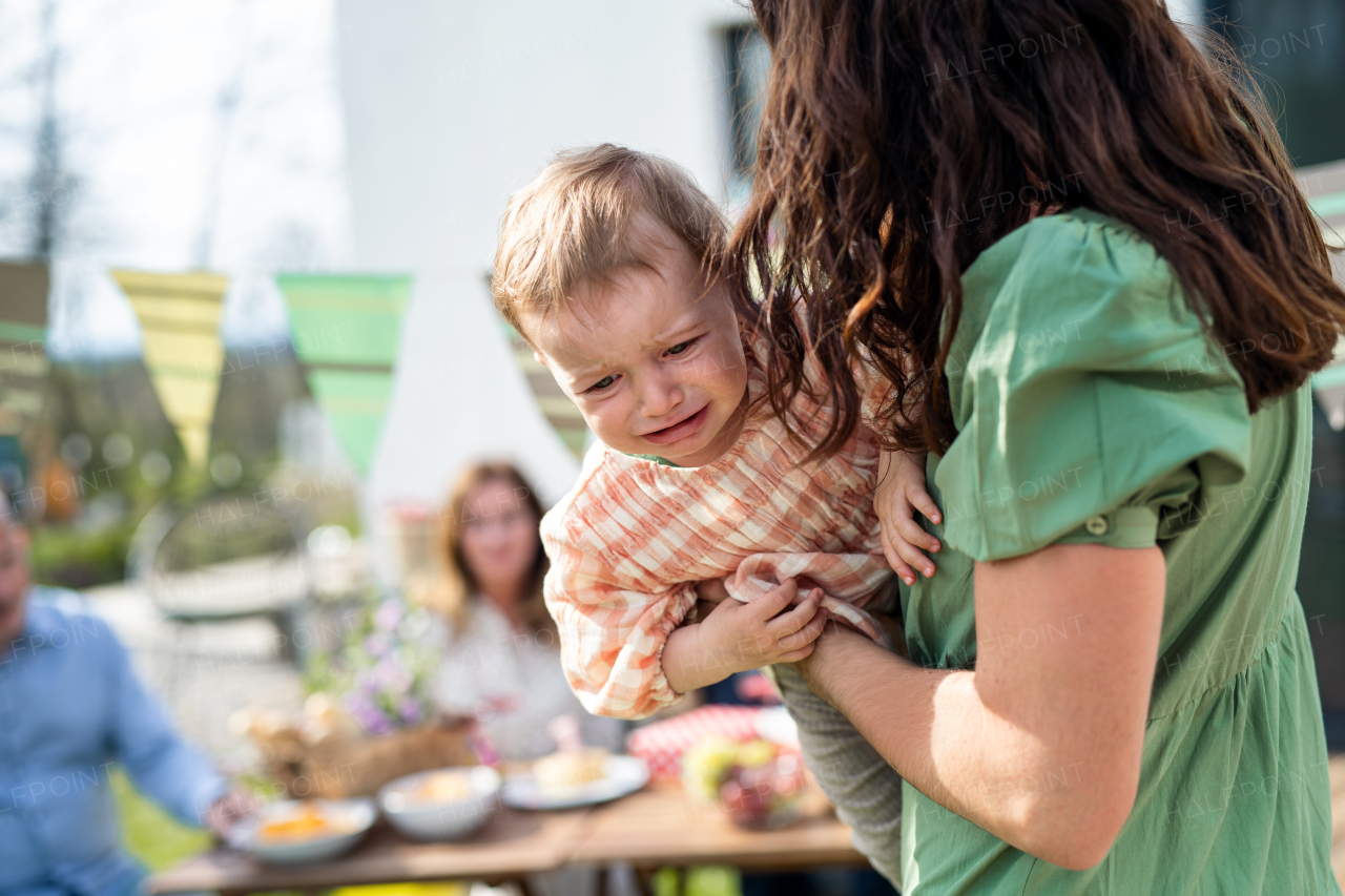 Upset crying baby with mother outdoors in garden at home, birthday celebration party.