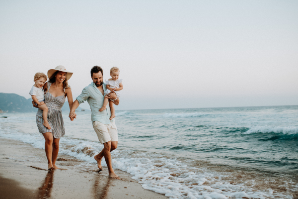 A family with two toddler children walking on beach on summer holiday at sunset. A father and mother carrying son and daughter in the arms.