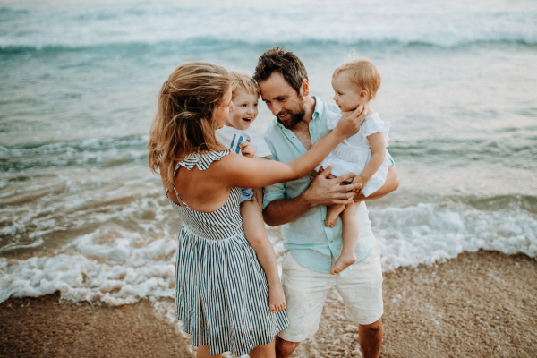 A happy young family with two toddler children standing on beach on summer holiday.