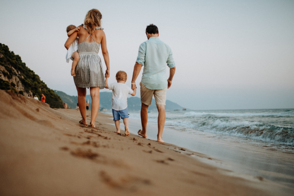 A rear view of young family with two toddler children walking on beach on summer holiday.