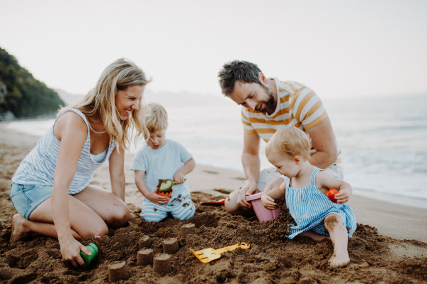 A young family with toddler children playing with sand on beach on summer holiday.