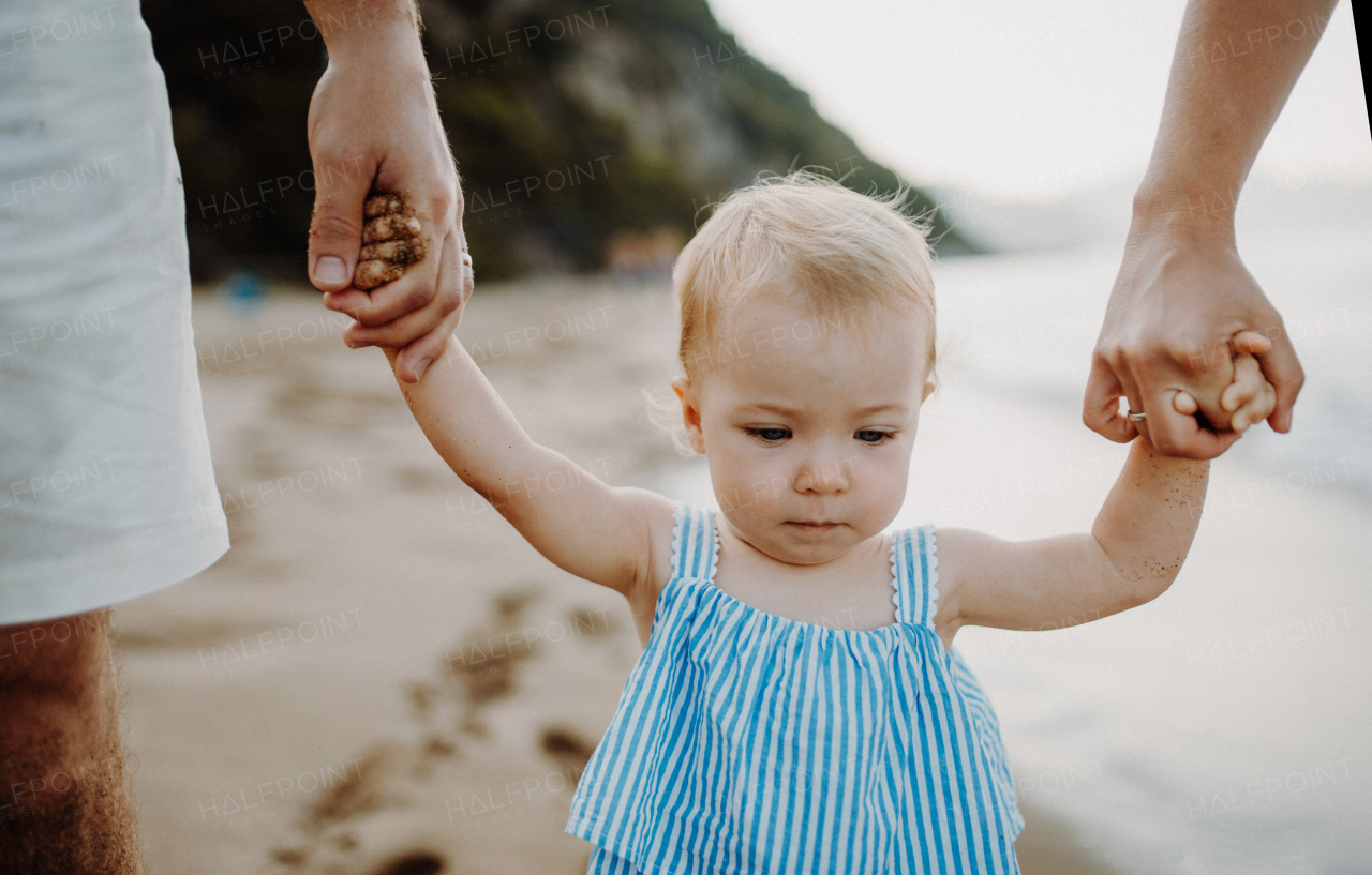 A midsection of parents with toddler daughter walking on beach on summer holiday, holding hands.