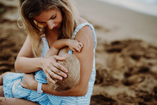 A young mother breasfeeding toddler daughter on beach on summer holiday.