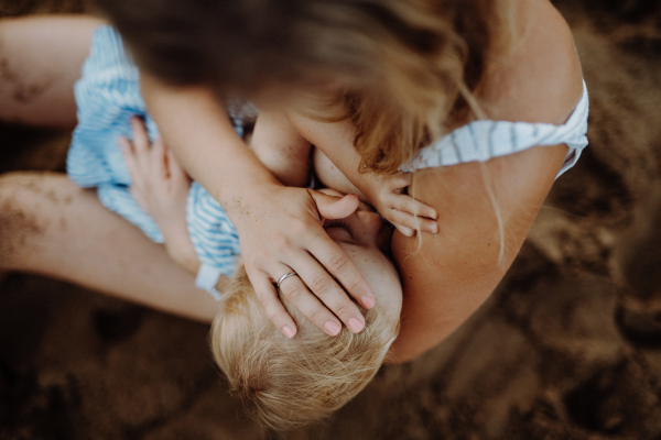 An unrecognizable young mother breasfeeding toddler daughter on beach on summer holiday. A top view.