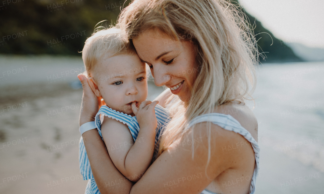 A young mother with small toddler daughter standing on beach on summer holiday, hugging.