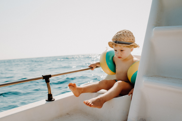 A small boy with armbands sitting on boat on summer holiday, holding a railing.