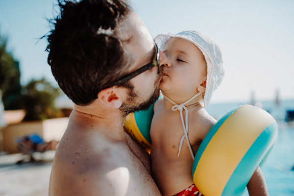 A father with small child with armbands standing by swimming pool on summer holiday.