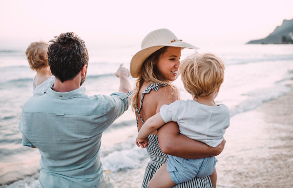 A family with two toddler children walking on beach on summer holiday at sunset. A father and mother carrying son and daughter in the arms.