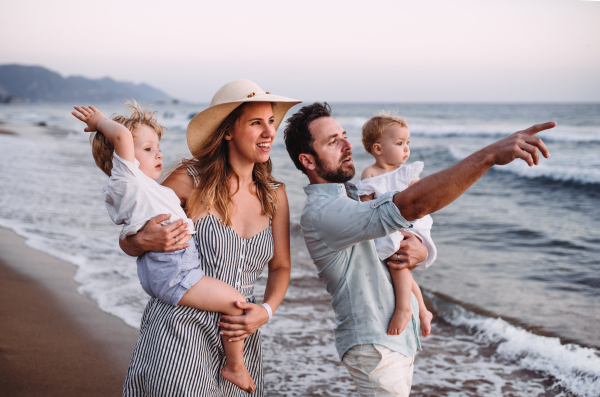 A family with two toddler children walking on beach on summer holiday at sunset. A father and mother carrying son and daughter in the arms.