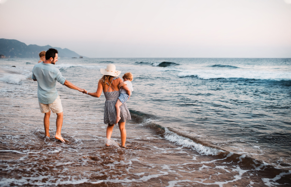 A rear view of young family with two toddler children walking on beach on summer holiday.