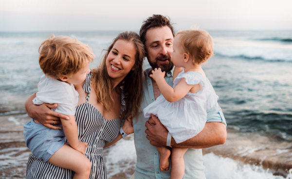 A young family with two toddler children standing on beach on summer holiday, laughing.