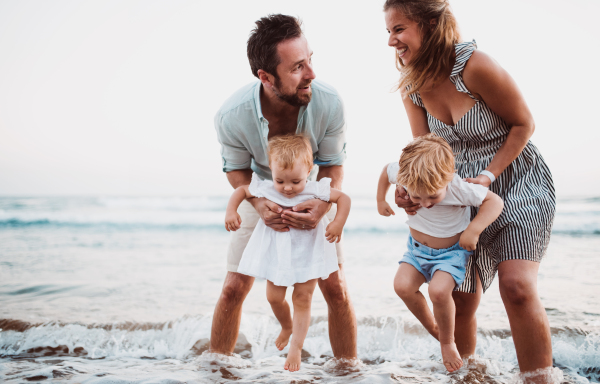 A young family with two toddler children standing on beach on summer holiday, having fun.