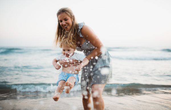 A young mother with a toddler boy standing on beach on summer holiday, having fun.