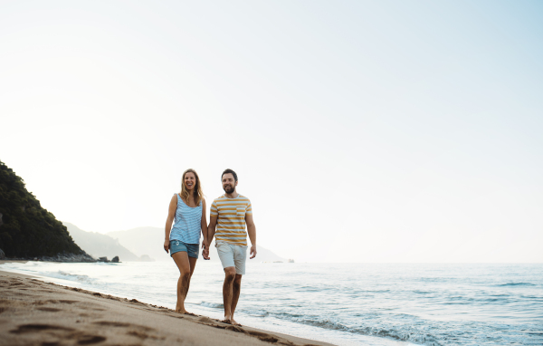 A cheerful man and woman walking on beach on summer holiday, holding hands. Copy space.