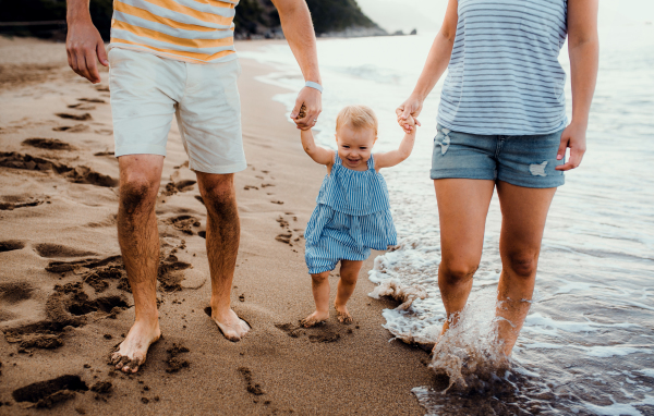 A midsection of parents with toddler daughter walking on beach on summer holiday, holding hands.