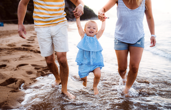 A midsection of parents with toddler daughter walking on beach on summer holiday, holding hands.