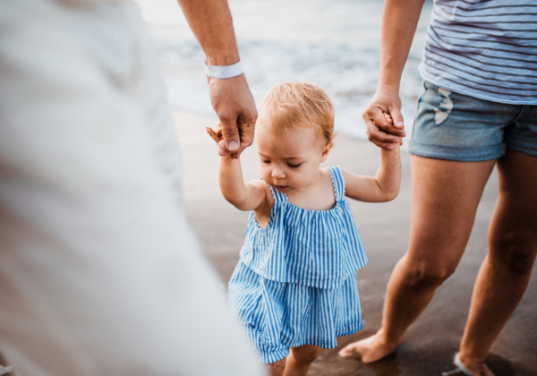 A midsection of parents with toddler daughter walking on beach on summer holiday, holding hands.