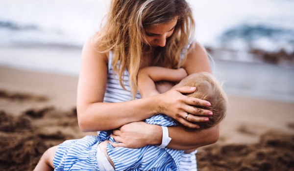 A young mother breasfeeding toddler daughter on beach on summer holiday.