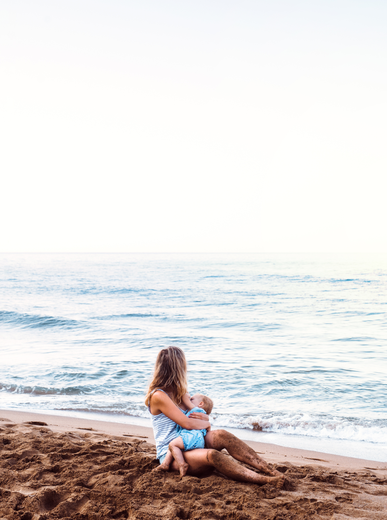 A young mother breasfeeding toddler daughter on beach on summer holiday.