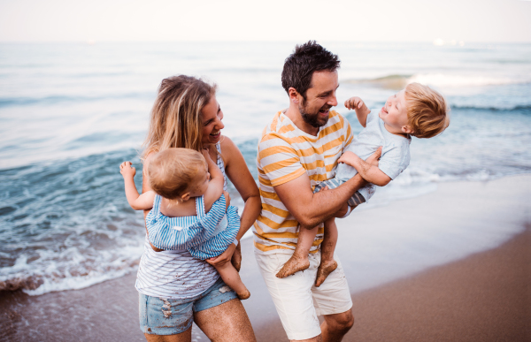 A young family with two toddler children walking on beach on summer holiday, laughing.