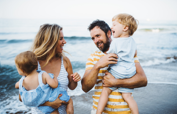 A young family with two toddler children standing on beach on summer holiday, laughing.