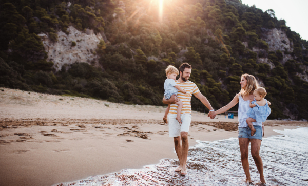 A family with two toddler children walking on beach on summer holiday at sunset. A father and mother carrying son and daughter in the arms, holding hands.